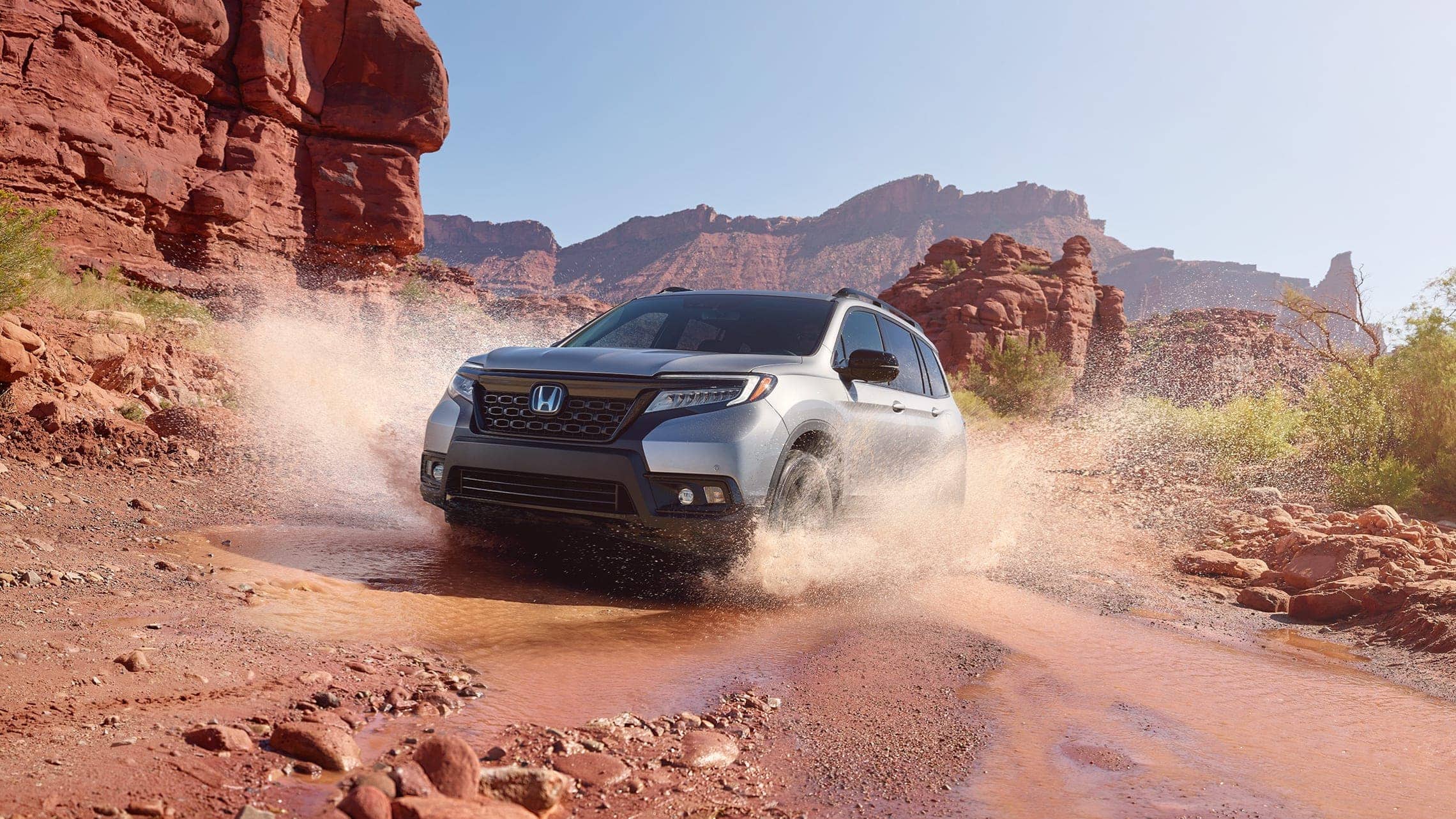 Driver-side front 3/4 view of 2019 Honda Passport Elite in Lunar Silver Metallic, demonstrating all-wheel drive and splashing through water on a rugged mountain road.