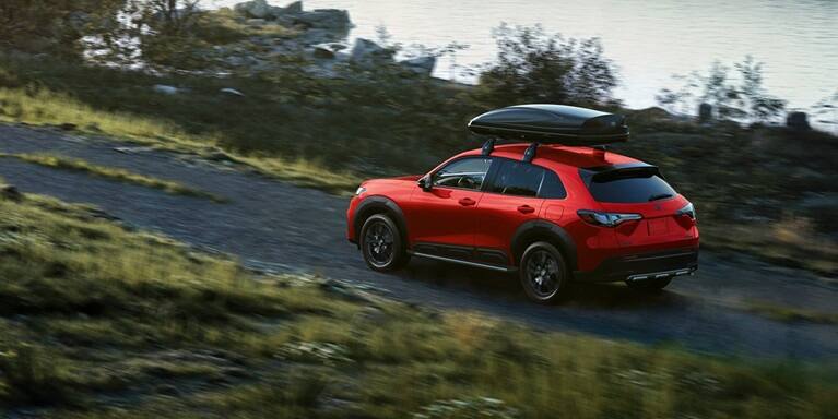 2027 Honda HR-V Sport in Milano Red profile view driving on a dirt road near water with a roof box (Subcompact Crossover SUV).
